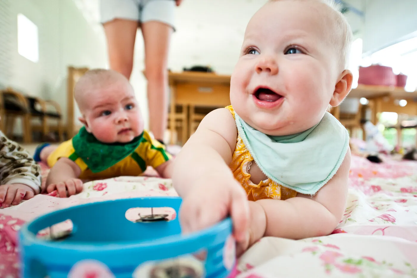 En glad baby der smiler og leger med en blå tamburin på et blomstret tæppe.